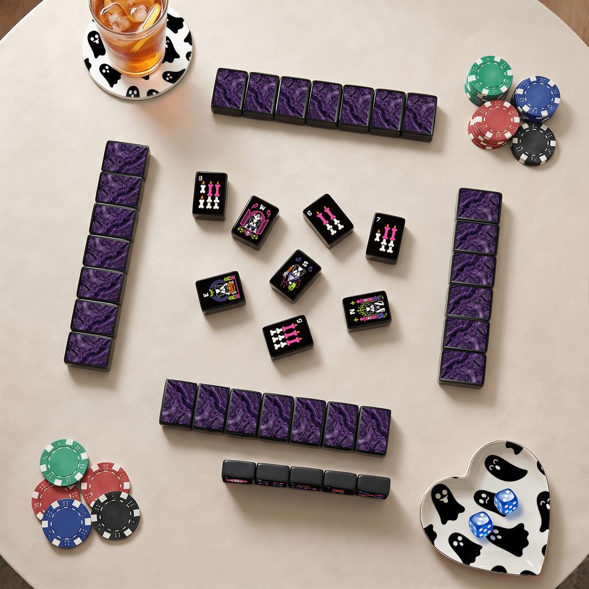 Halloween mahjong game night flat lay – purple textured tile walls, black bulldog tiles, colorful poker chips, ghost-print heart bowl, and an iced cocktail on a beige table