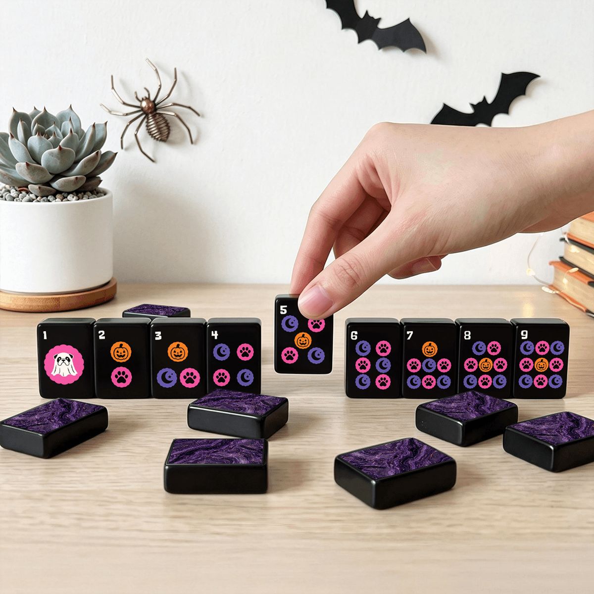 Hand placing a Halloween mahjong dots tile on a wood table with purple marbled tile backs, succulent, and bat decorations – spooky game night lifestyle scene with pink ghost and pumpkin symbols