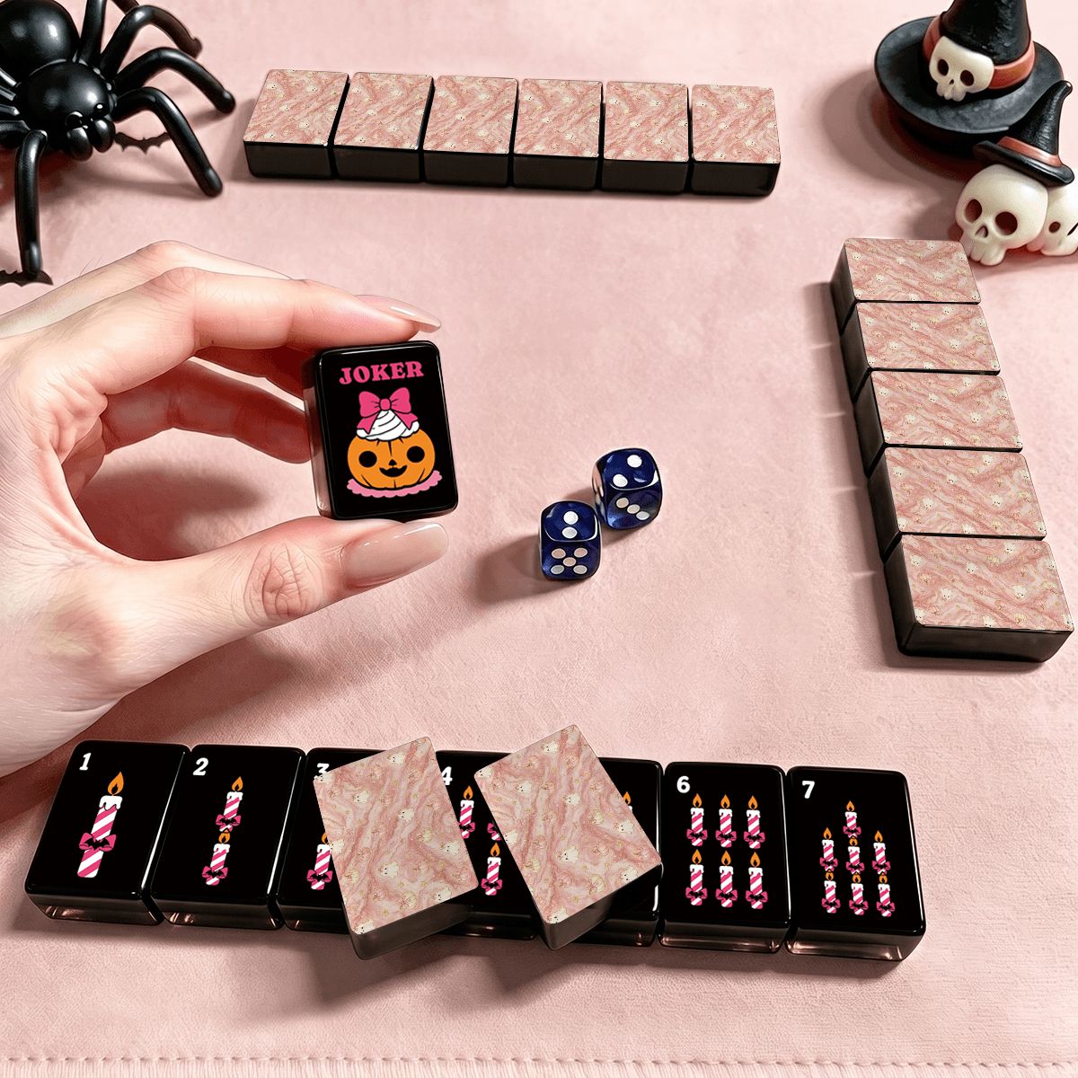 Hand holding a Gothic Dessert Halloween mahjong Joker tile with pink pumpkin cupcake and bow design, styled with blue dice, pink marble tile backs, spider prop, and skull witch hat on pink surface