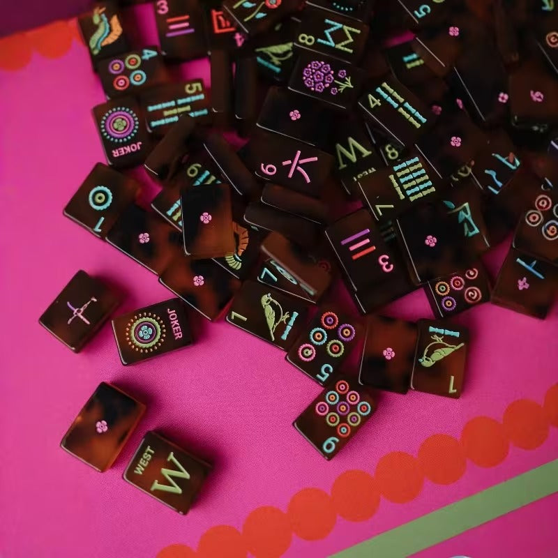 Overhead scatter of Amber Tortoiseshell American mahjong tiles with West wind (W) and Joker tiles – amber-brown marbled 160-tile set showing dots, bams, cracks, and flower tiles on pink tabletop