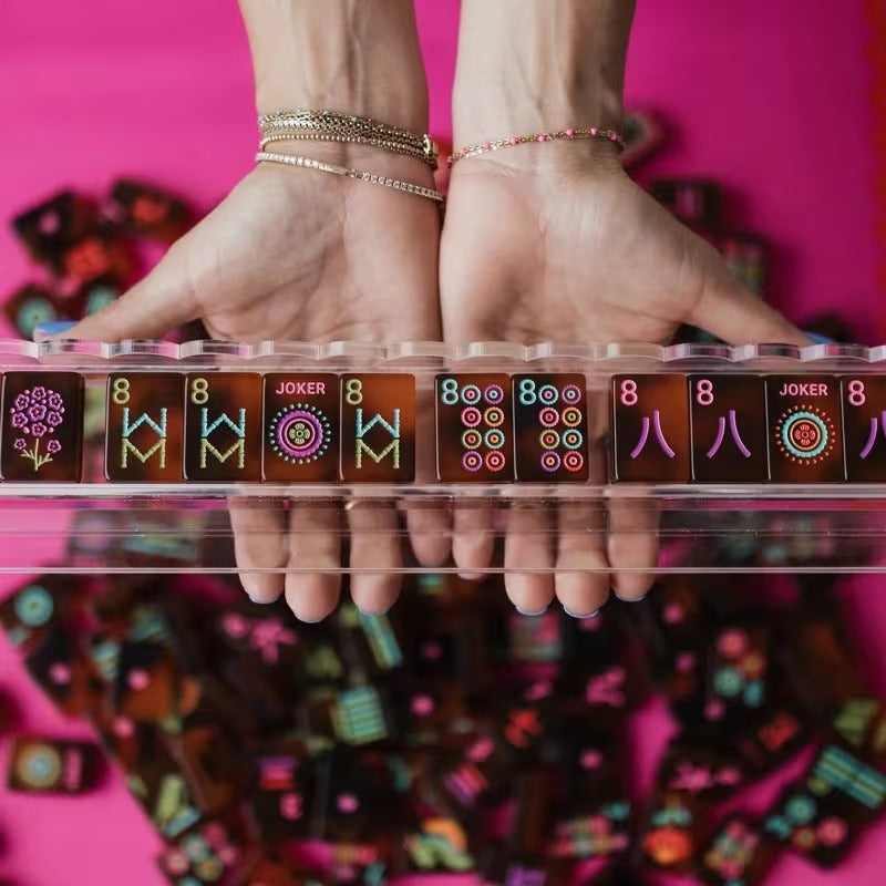Hands holding a clear mahjong rack with a row of amber tortoiseshell American mahjong tiles – flower, winds (W), Jokers, and 8 dots tiles visible in a colorful hand-painted style