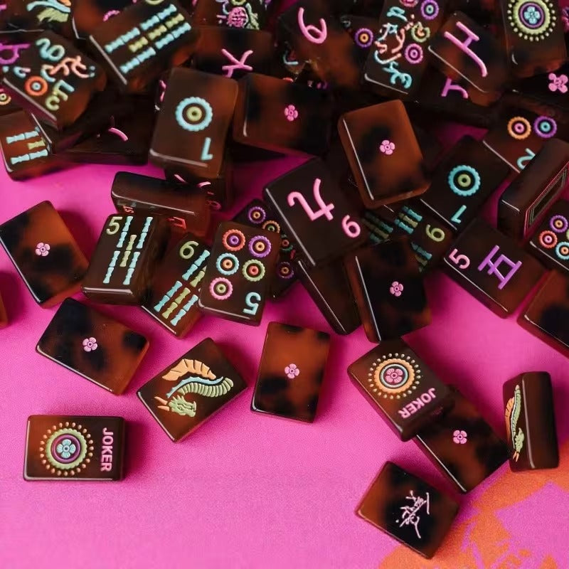 Scattered amber tortoiseshell mahjong tiles on a pink surface – close-up showing glossy amber-and-brown marbled tiles with colorful numerals, dots, bams, floral flower motifs, and pink Joker tiles