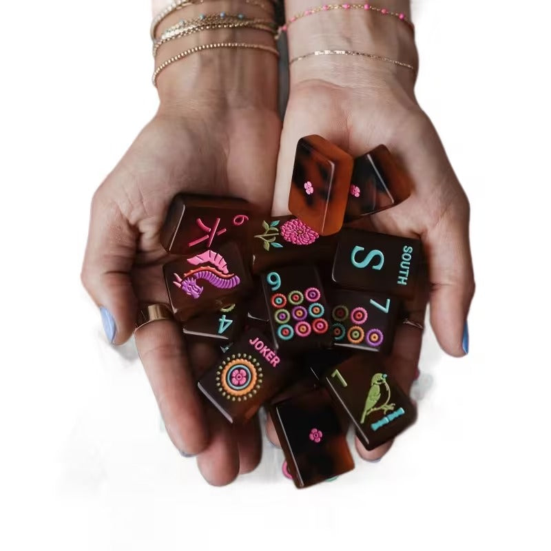 Hands cupping a handful of amber tortoiseshell American mahjong tiles on a clean white background – detailed view of South wind, 7 dots, 6 bams, Joker, and floral bird tiles with vivid painted colors