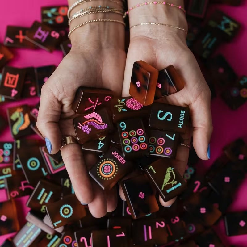Hands holding a pile of Amber Tortoiseshell American mahjong tiles – rich amber-brown tiles with colorful engraved dots, bams, cracks, joker and floral designs on a pink background